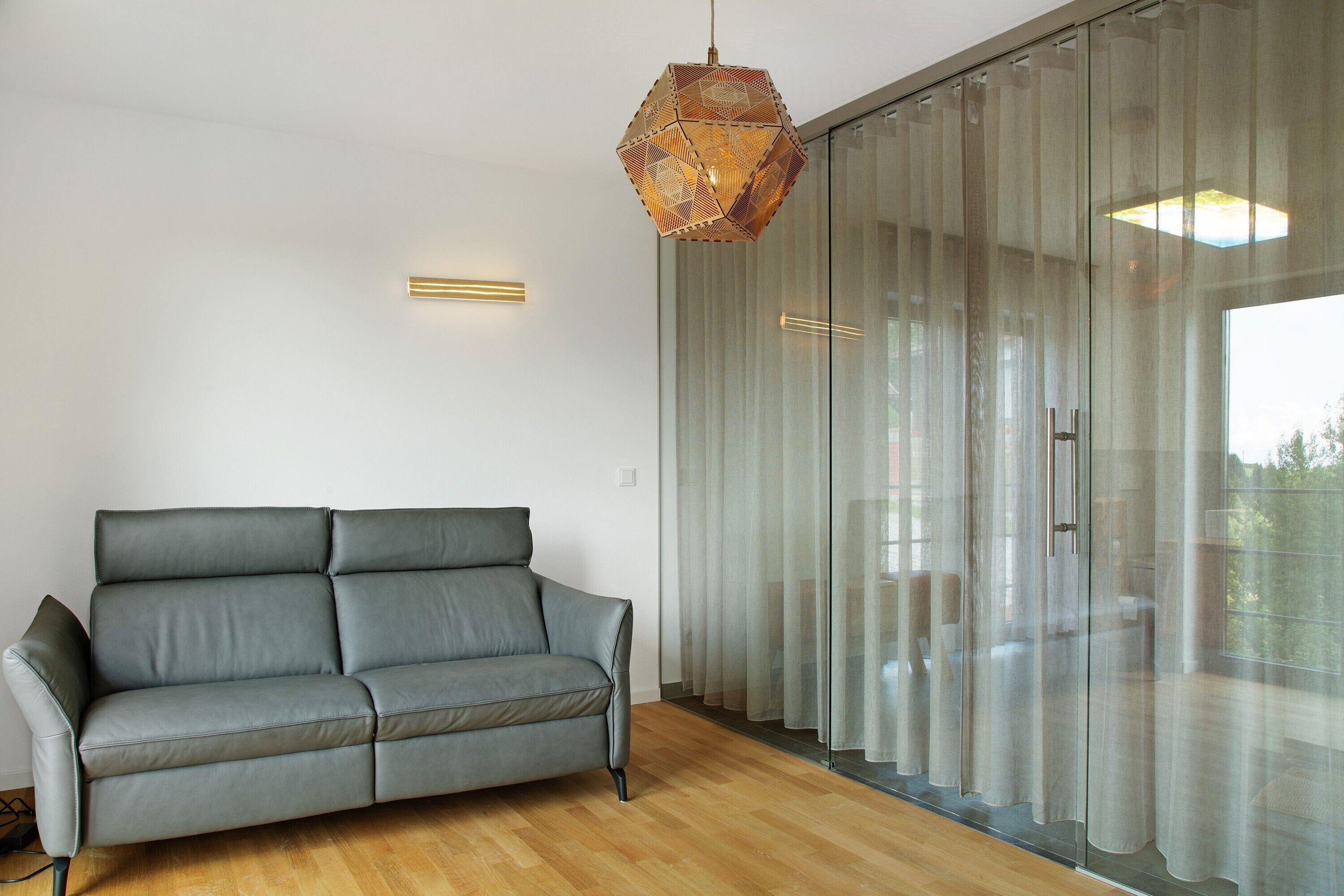 Un salon moderne avec un canapé gris, un luminaire suspendu aux formes géométriques et une cloison en verre agrémentée de rideaux.