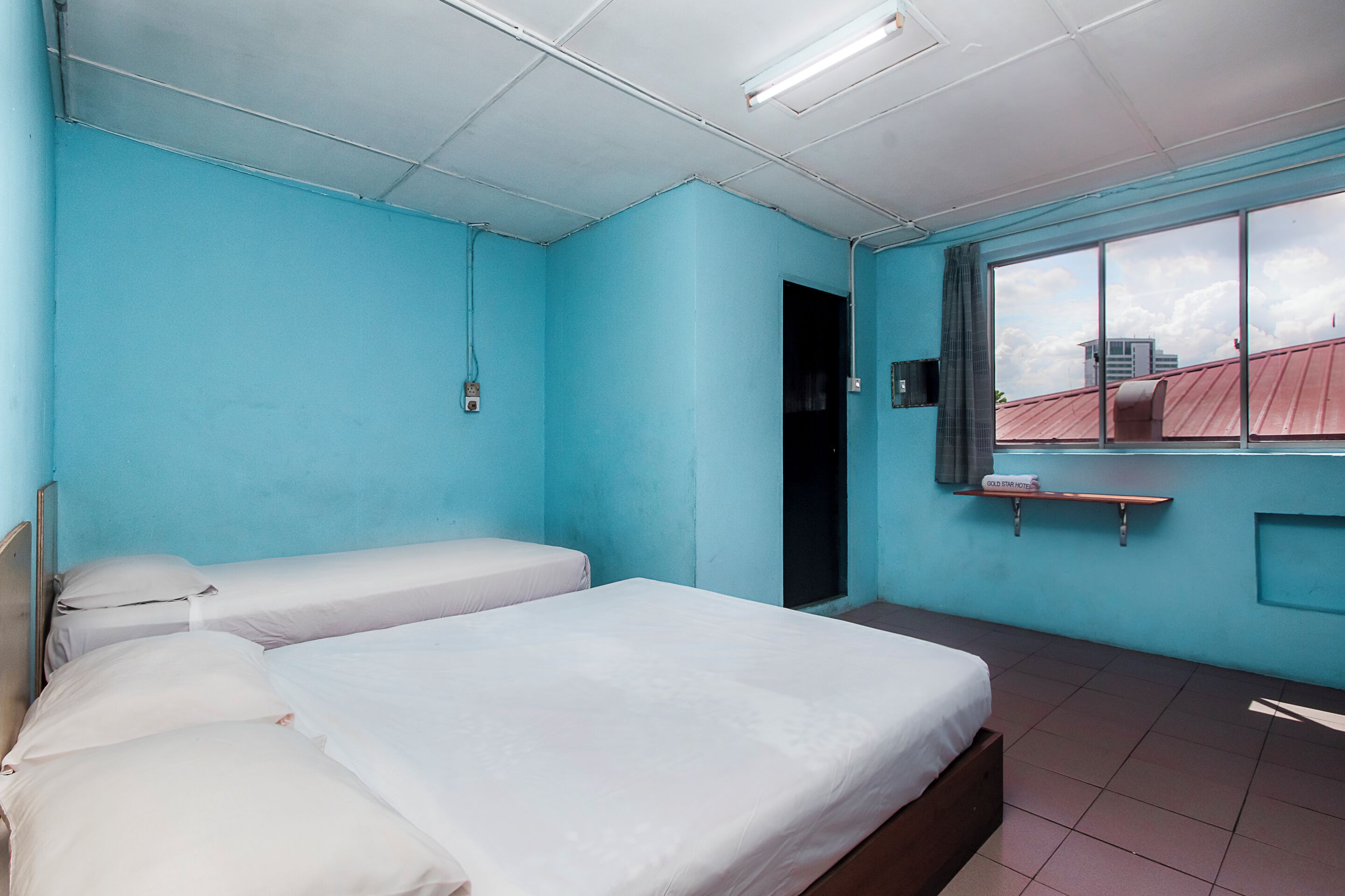 Una habitación con dos camas, una ventana con vistas a edificios, un estante con una toalla y una puerta.