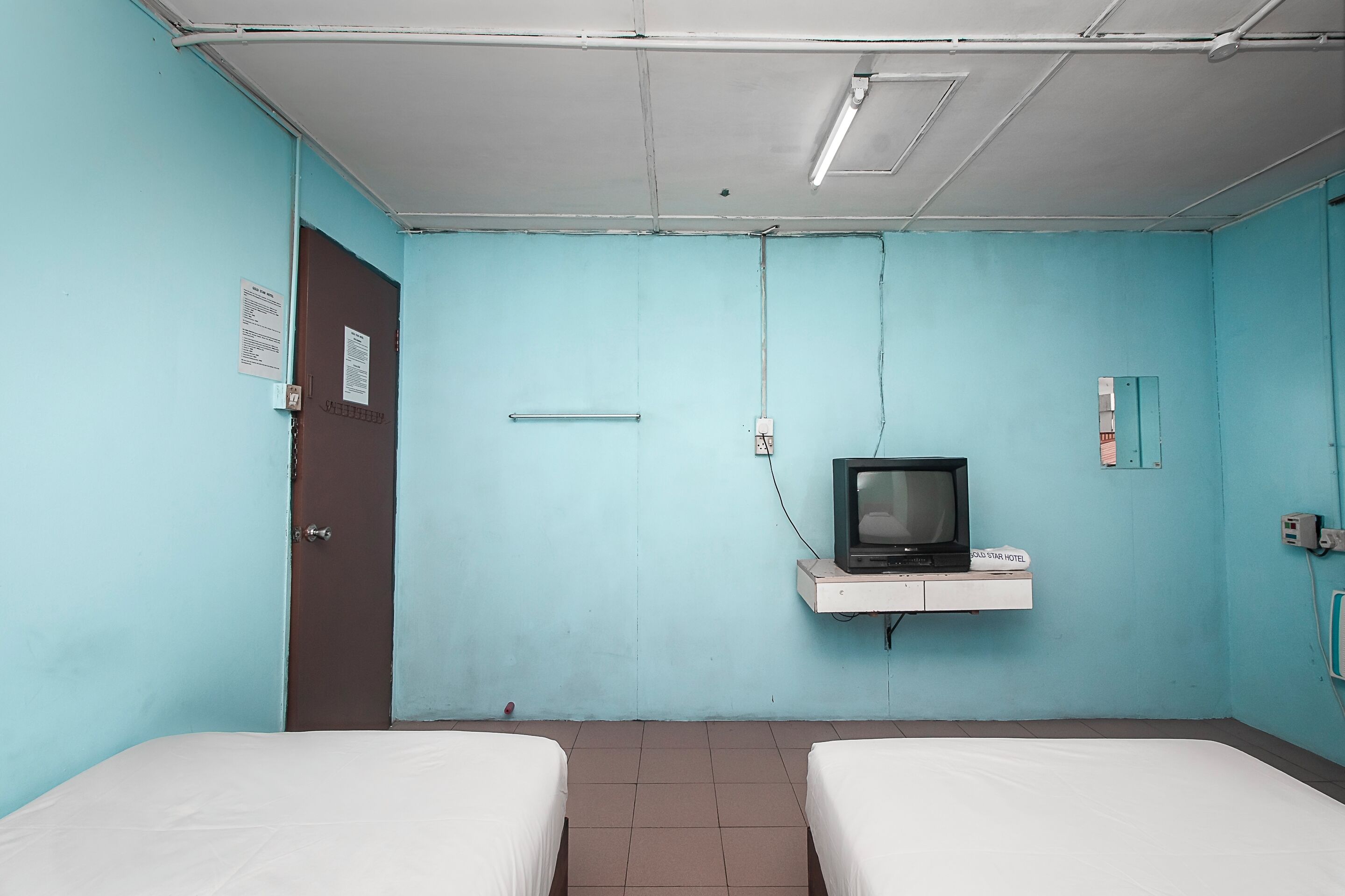 Una habitación con dos camas, un televisor en un estante, una puerta y un interruptor de luz en la pared.