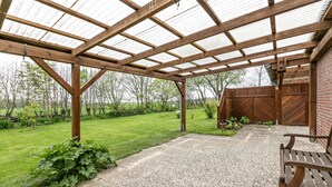 Un patio abrité avec un sol en gravier, des bancs en bois et une vue sur une pelouse verdoyante et des arbres.