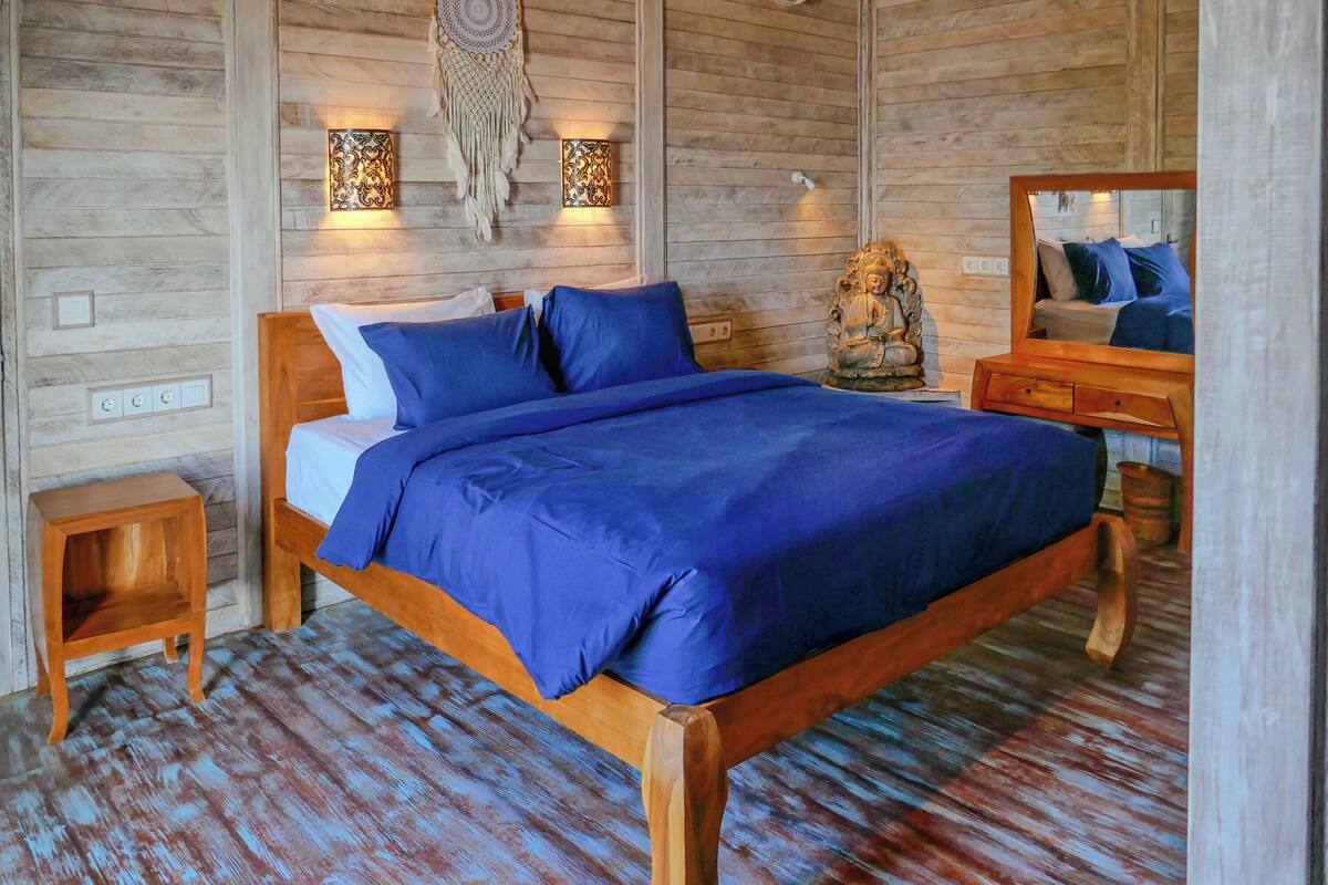 Ein Bett mit Holzrahmen, dazu blaue Bettwäsche, ein kleiner Nachttisch aus Holz und eine Wandlampe.
