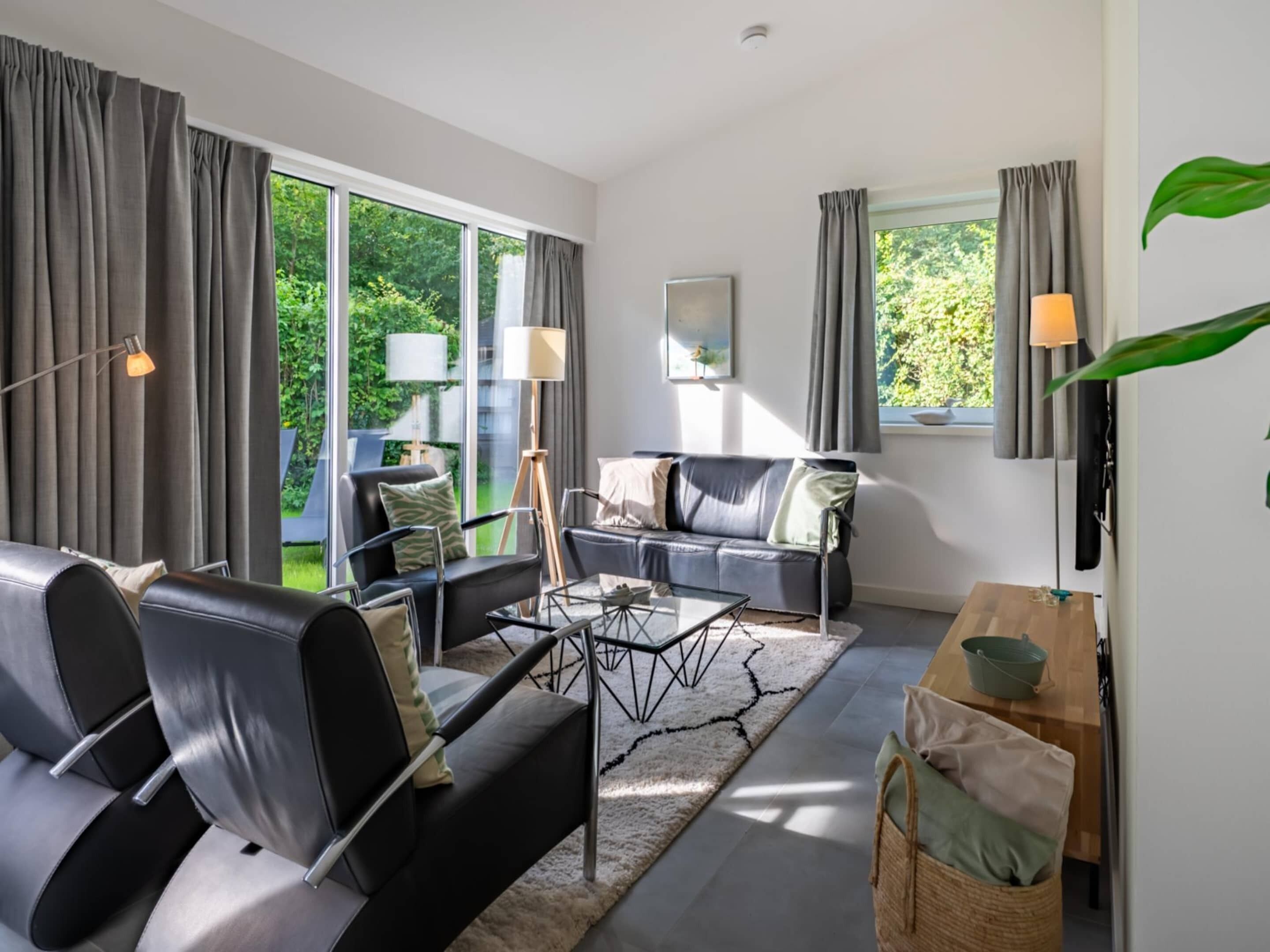 Un salon moderne avec un canapé, des fauteuils, une table basse et une vue sur la verdure grâce à de grandes fenêtres.