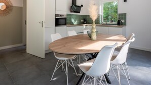 Eine moderne Küche mit weißen Schränken, einem Holzespeisetisch und weißen Stühlen. Auf dem Tisch steht eine Vase mit Trockenpflanzen.
