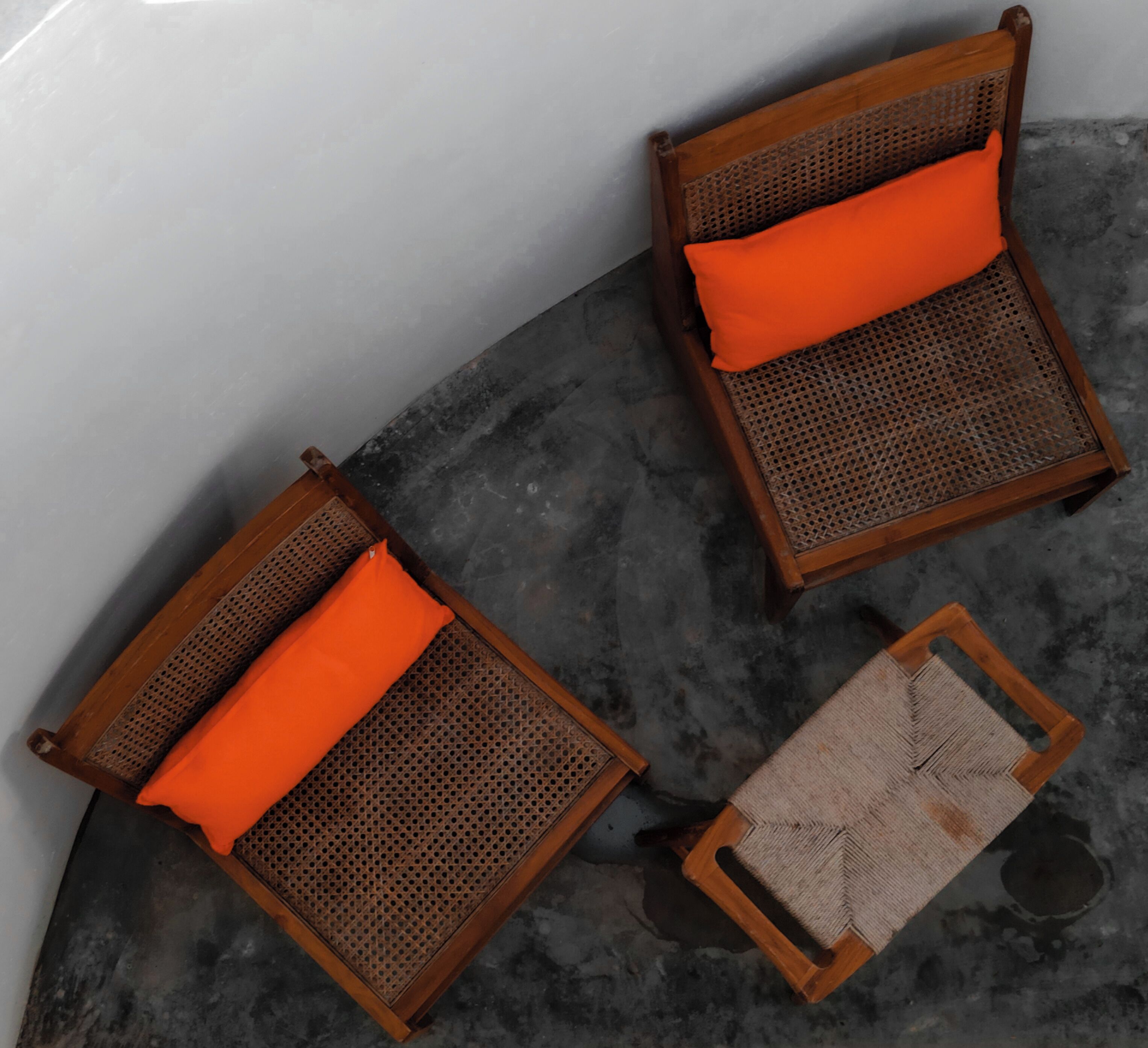 Dos sillas de mimbre con cojines naranja y una pequeña mesa de madera con asiento tejido, sobre un suelo circular de hormigón.