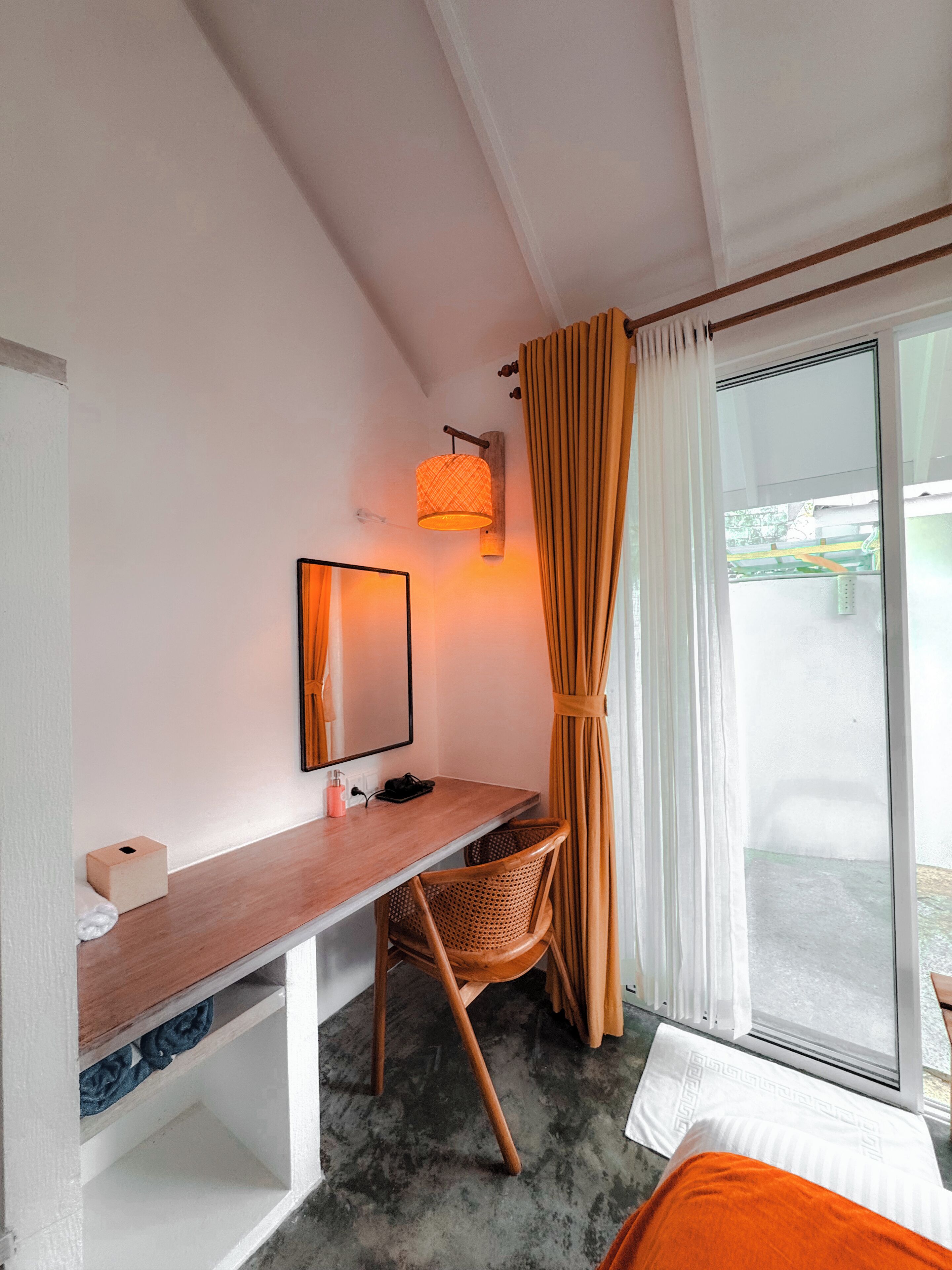 A room with a wooden desk, a chair, a bed, a window with curtains, and a wall-mounted lamp.