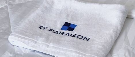 A folded white towel with the text "D'PARAGON" and a blue logo on a bed with striped bedding.