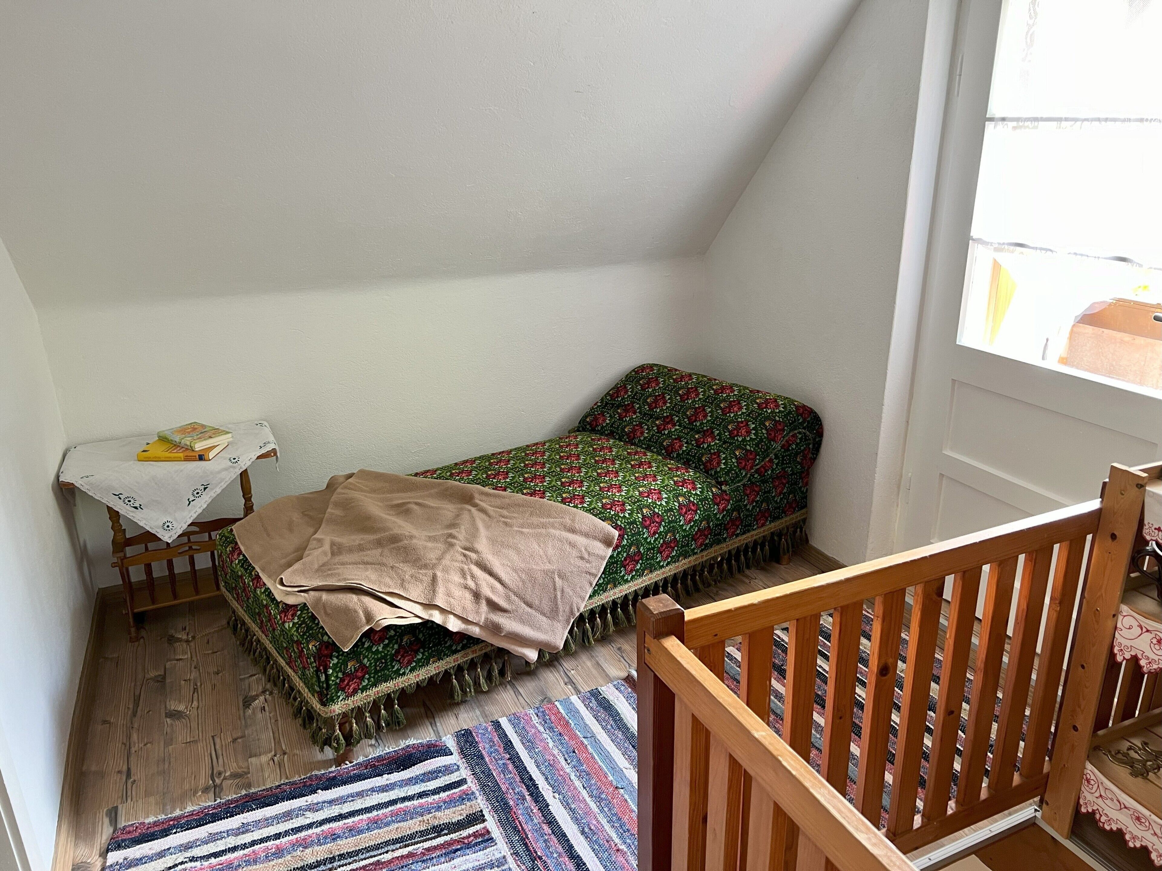 A small, cozy room with a single bed, a small table with books, and a wooden chair.