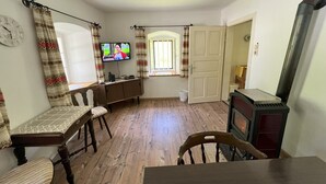 Una habitación con piso de madera, una mesa de comedor con sillas, un televisor montado en la pared, una ventana con cortinas y una puerta que da a otra habitación.