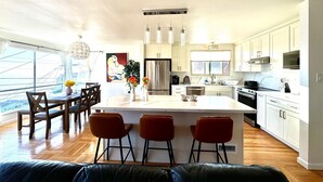 A modern kitchen with white cabinets, a central island with bar stools, a refrigerator, stove, microwave, and a large window providing natural light. - Spacious 4BR Ocean‑View Home • Modern Kitchen • Near Beach & SF (Daly City)