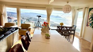 A spacious living area with a large dining table, comfortable seating, and a view of the cityscape through large windows. - Spacious 4BR Ocean‑View Home • Modern Kitchen • Near Beach & SF (Daly City)