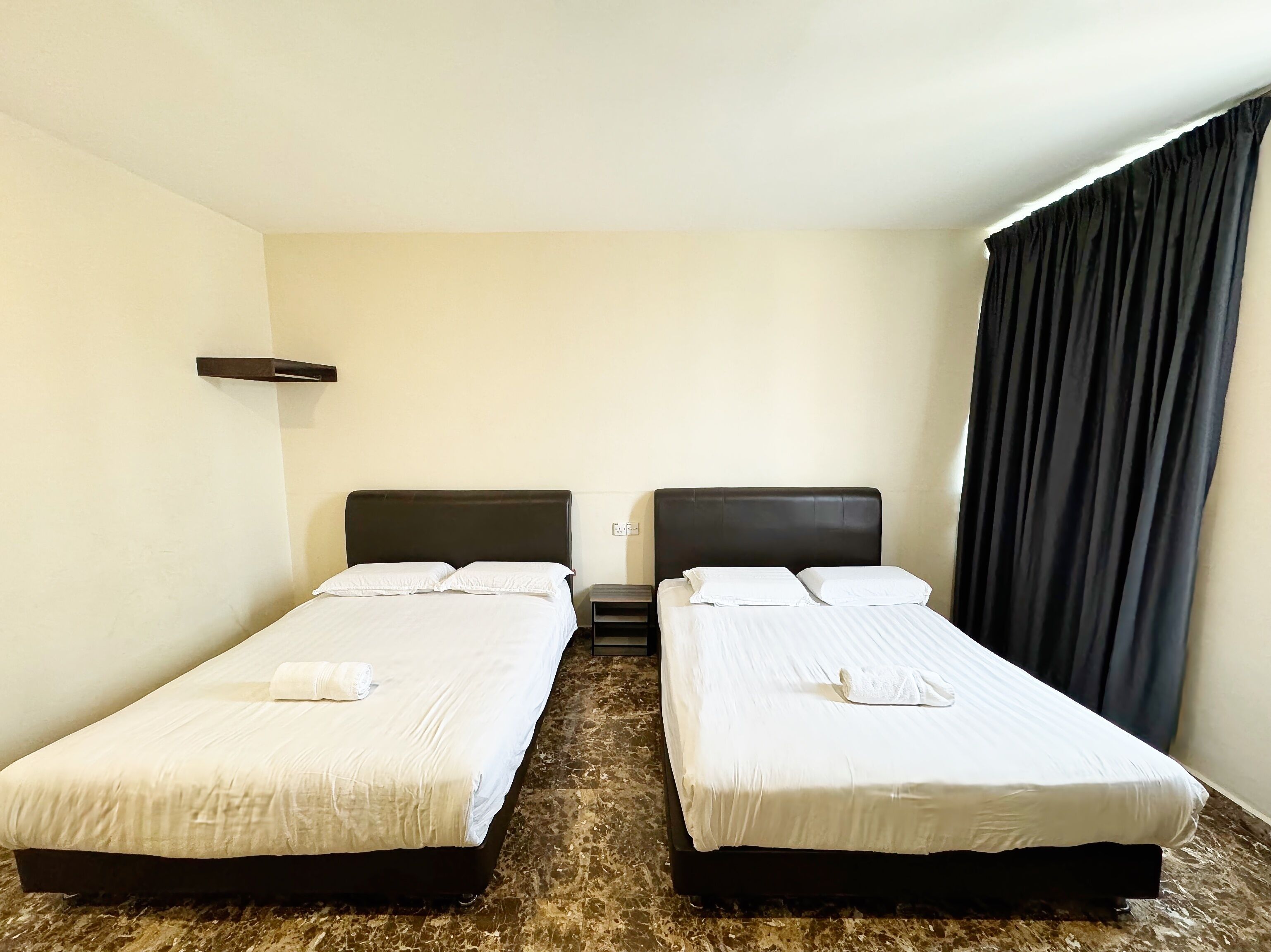 Una habitación de hotel con dos camas individuales, un estante pequeño, suelo de mármol y una cortina oscura.