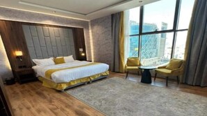 Habitación de hotel moderna con una cama grande, una mesita, dos sillas y vistas a la ciudad a través de amplios ventanales.