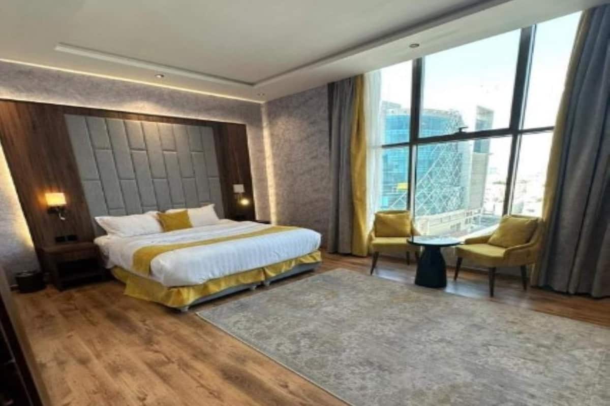 A modern hotel room with a large bed, a small table, two chairs, and a view of the city through large windows.
