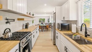 A modern kitchen with white cabinets, a wooden countertop, stainless steel appliances, and a dining area in the background.