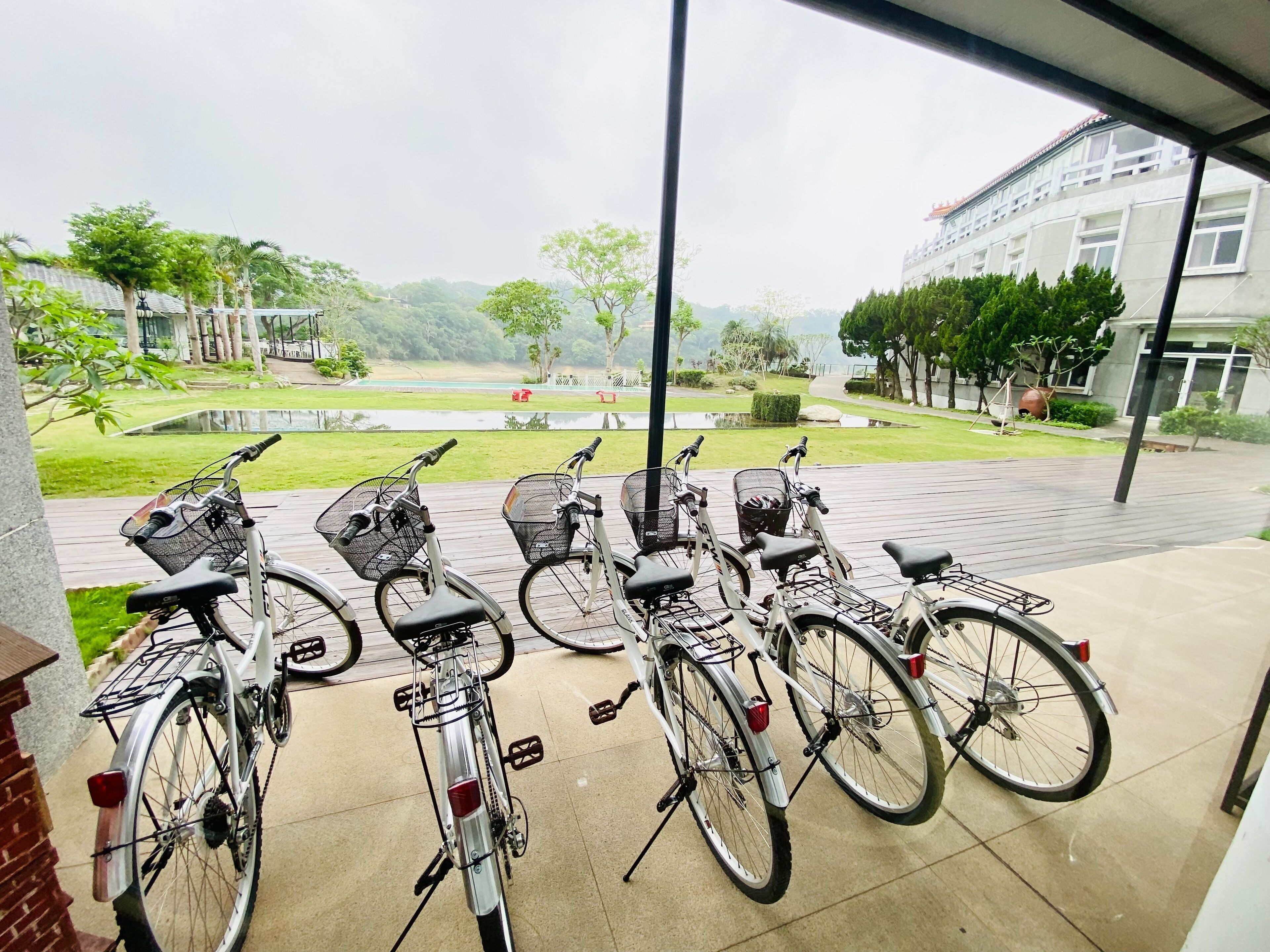 A row of bicycles parked on a covered outdoor area with a view of a landscaped garden and a building in the background.