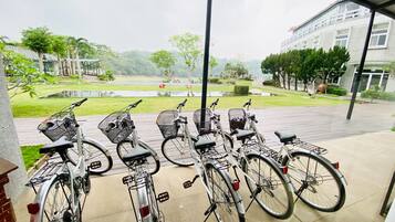 A row of bicycles parked on a covered outdoor area with a view of a landscaped garden and a building in the background.