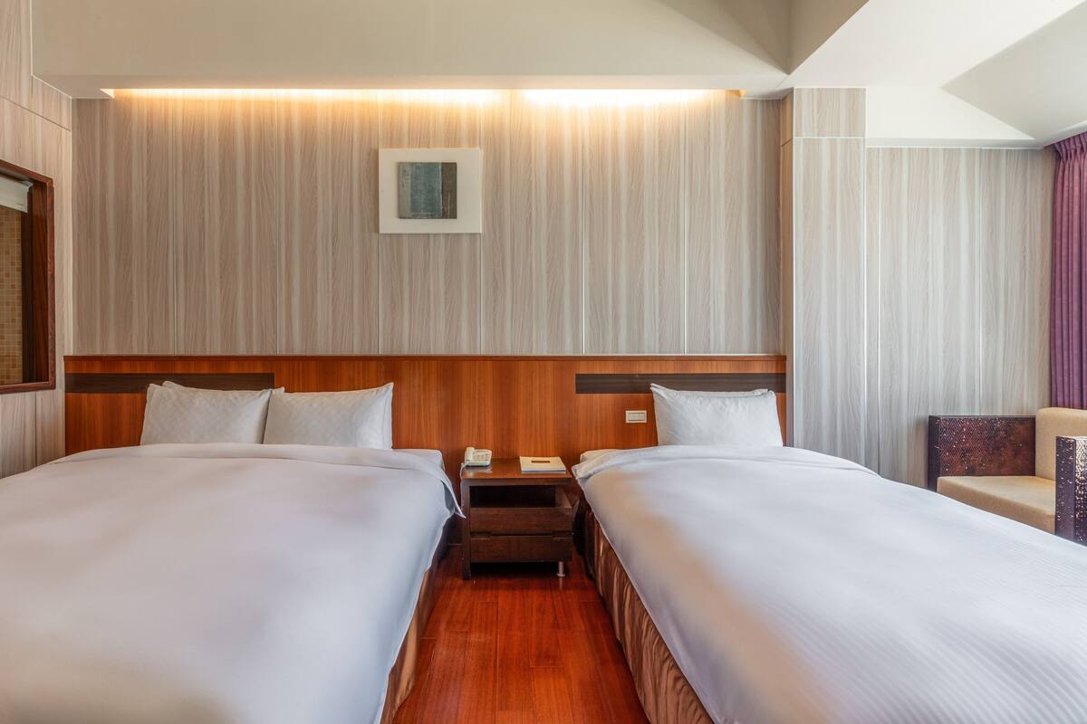 Habitación de hotel con dos camas, cabecero de madera, mesitas de noche, un pequeño escritorio y una silla.