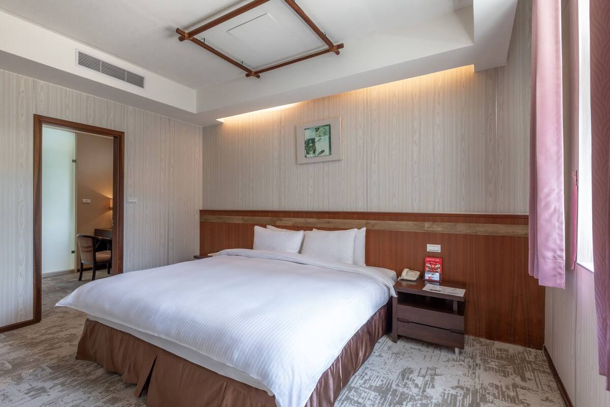Habitación de hotel con una cama grande, cabecera de madera, una mesita de noche, un cuadro enmarcado en la pared y una puerta que da a otra habitación.