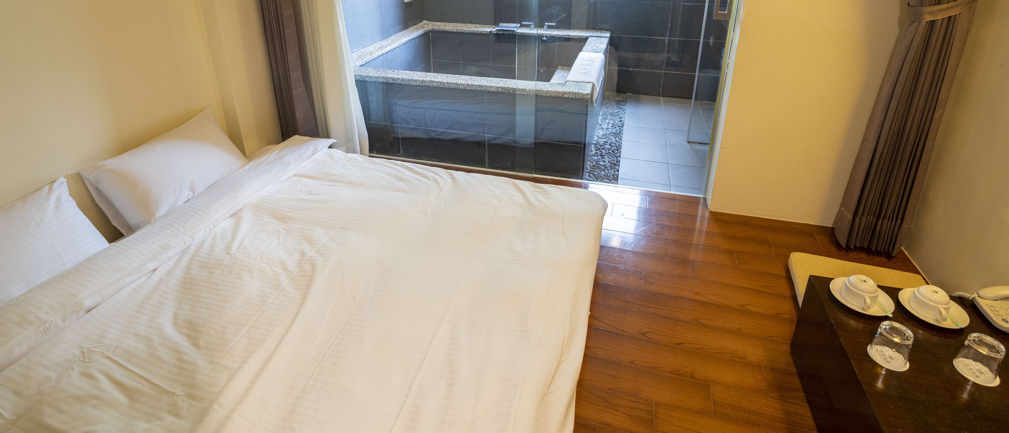 A hotel room with a large bed, a wooden floor, a glass shower enclosure, and a table with a telephone and glasses.