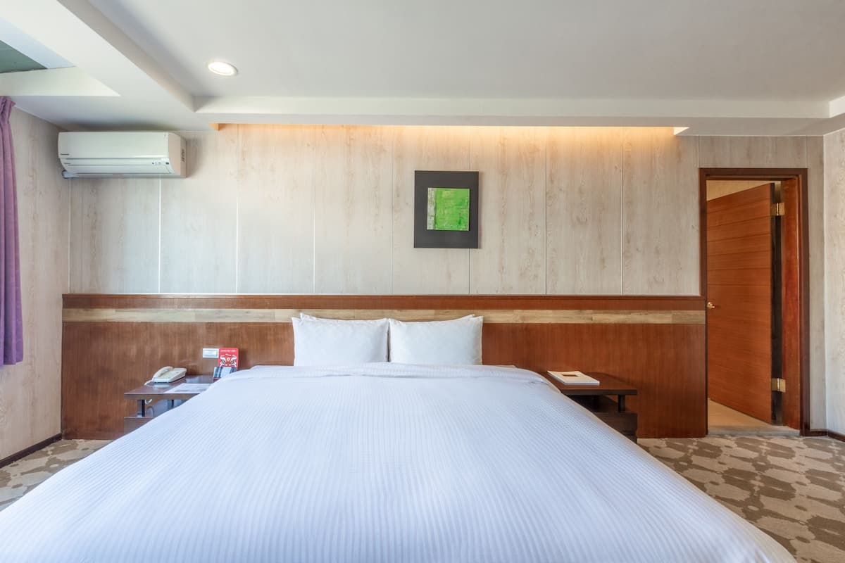 Habitación de hotel con una cama grande, cabecero de madera, un cuadro enmarcado en la pared, una mesita de noche con un teléfono y un equipo de aire acondicionado.