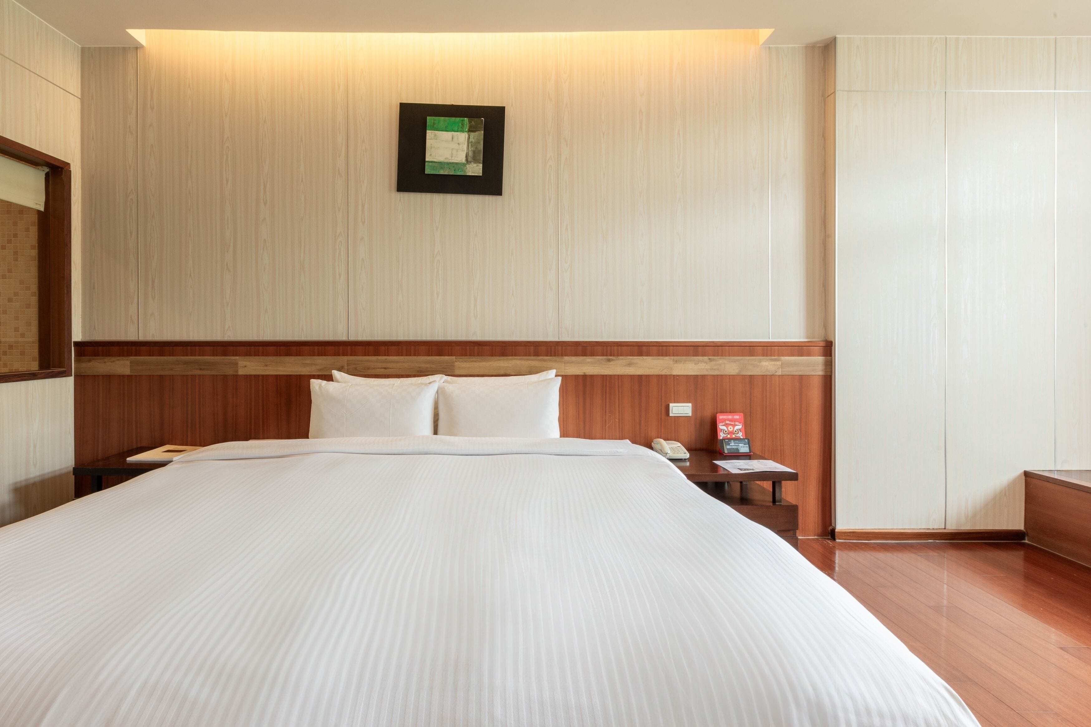 A hotel room with a large bed, wooden headboard, a bedside table with a phone and a red object, and a framed picture on the wall.
