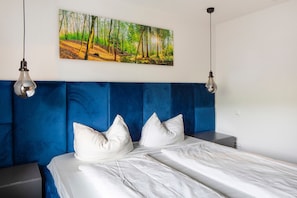 A bedroom with a blue headboard, white bedding, and a forest landscape painting above the bed.