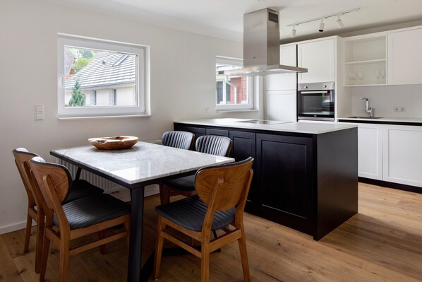 A modern kitchen with a central island, wooden chairs, a marble table, and white cabinetry.