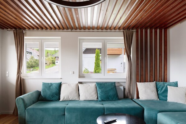 Un canapé de couleur bleu sarcelle agrémenté de coussins blancs et bleu sarcelle, un plafond en bois avec des lambris et une fenêtre avec des rideaux.
