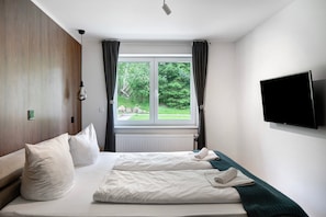 Una habitación de hotel con cama, ventana con cortinas, televisor de pantalla plana en la pared y termostato en la pared.