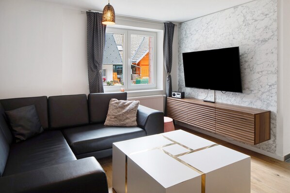 Un salon moderne avec un canapé d’angle, une table basse blanche, un meuble TV en bois et une grande fenêtre agrémentée de rideaux.