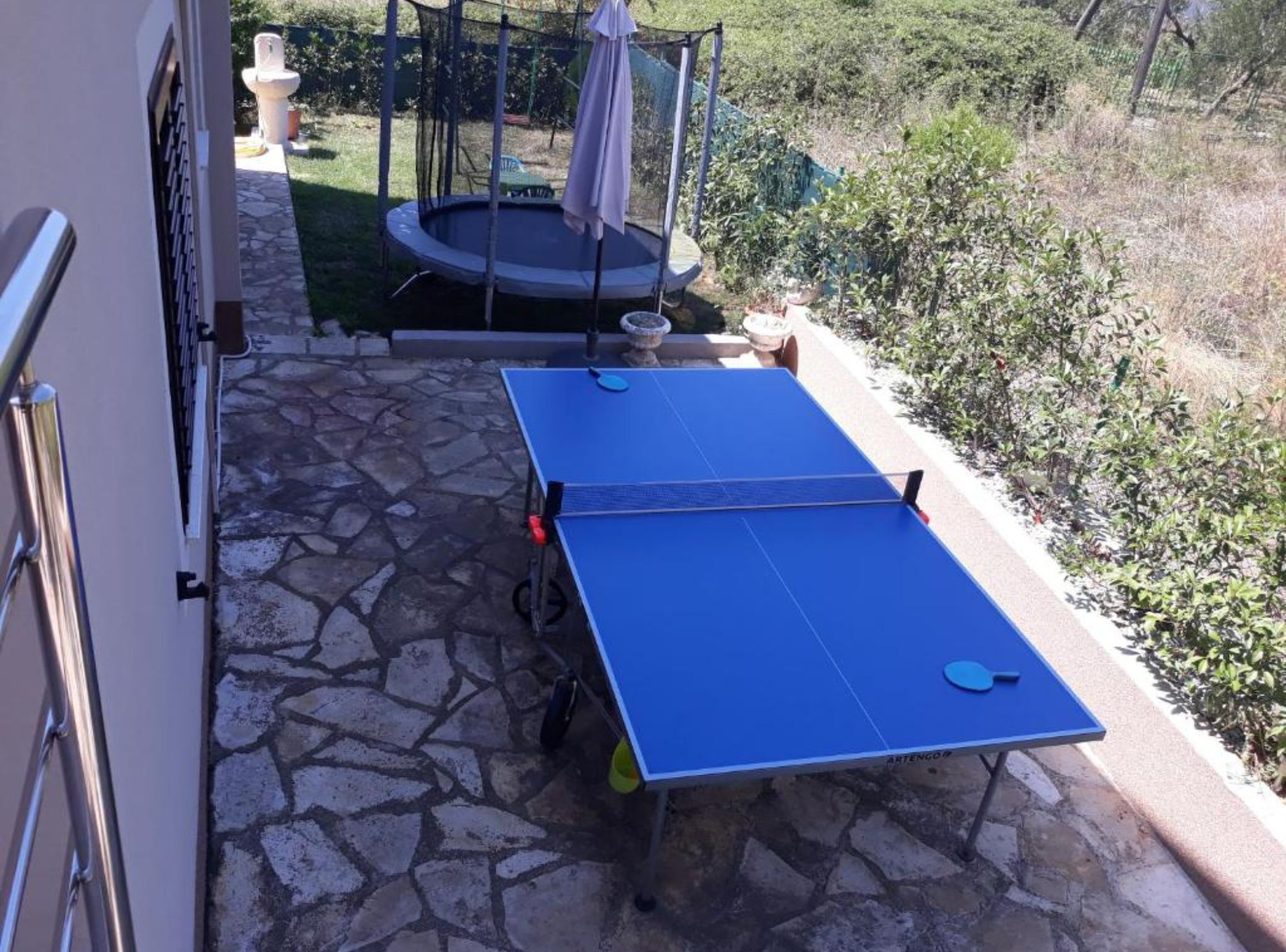 Mesa de ping pong instalada al aire libre, con un patio de piedra y un trampolín al fondo.