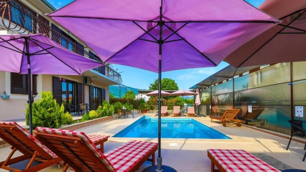 A poolside area with lounge chairs and umbrellas, a clear blue pool, and a building with a balcony in the background. - Villa Marica (Kastela)