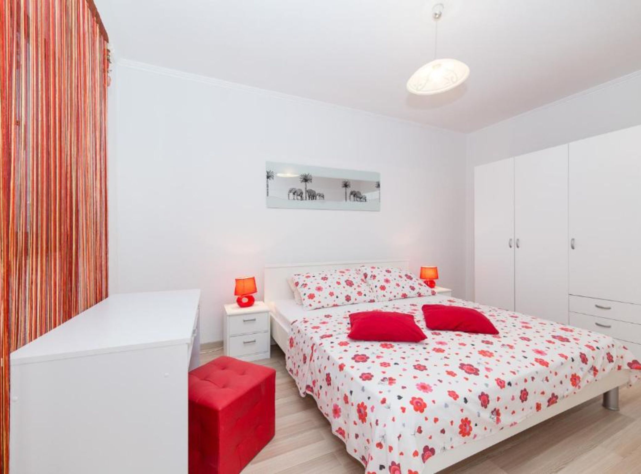 Un dormitorio con cama, dos mesitas de noche, un reposapiés rojo, un armario y un espejo fijado a la pared.