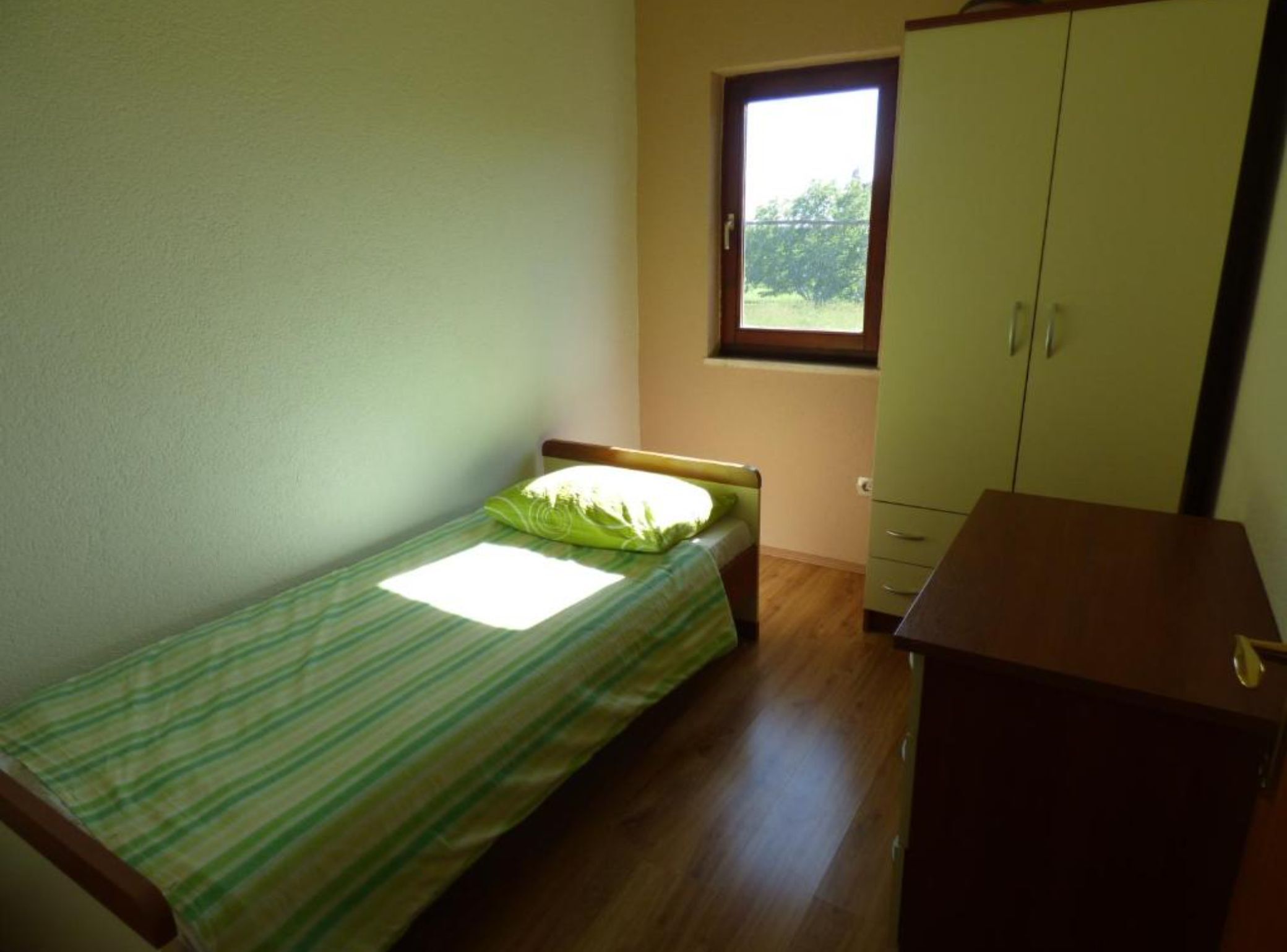 A small, single-bed room with a window, a wooden wardrobe, and a green-striped bedspread.