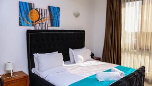 A neatly made bed with white bedding and a teal accent, a wooden nightstand with a lamp, and abstract art on the wall. - Myra Residences Kilimani (Nairobi)