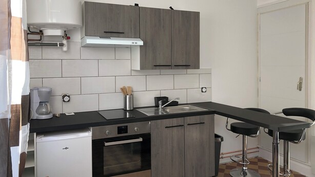 Een kleine keuken met grijze kastjes, een zwart aanrecht, een ingebouwde oven, een koffiezetapparaat en twee barkrukken.