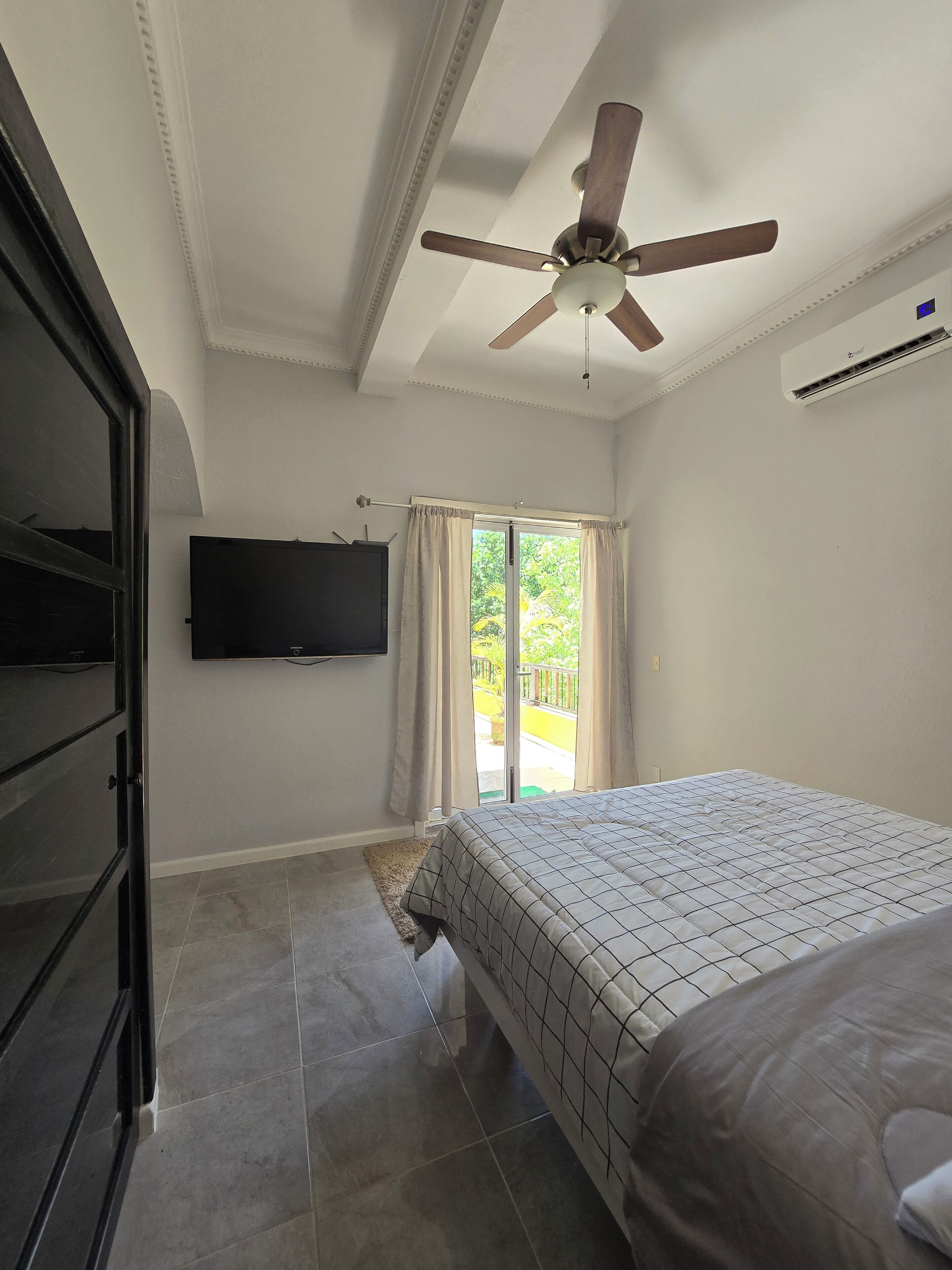 Ein Schlafzimmer mit einem Bett, einem Deckenventilator, einem an der Wand montierten Fernseher und einer Glasschiebetür, die zu einem Außenbereich führt.