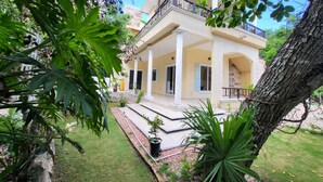 A two-story building with a balcony, columns, and a garden area with plants and a tree.