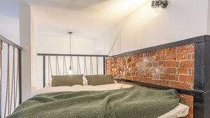 Une chambre avec une tête de lit en briques, un lit recouvert d’une couverture verte et un luminaire suspendu.