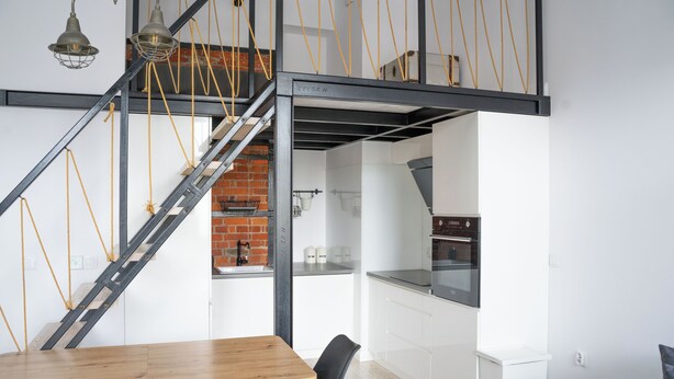 Une cuisine moderne avec des meubles blancs et un mur en briques, une table à manger en bois avec des chaises, et un escalier avec des poutres apparentes et des cordes jaunes.