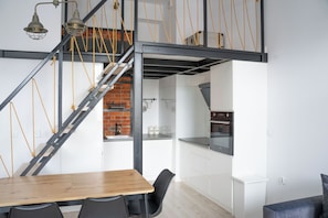 A modern kitchen with white cabinets and a brick wall, a wooden dining table with chairs, and a staircase with exposed beams and yellow ropes. - Nowa Papiernia (Wroclaw)