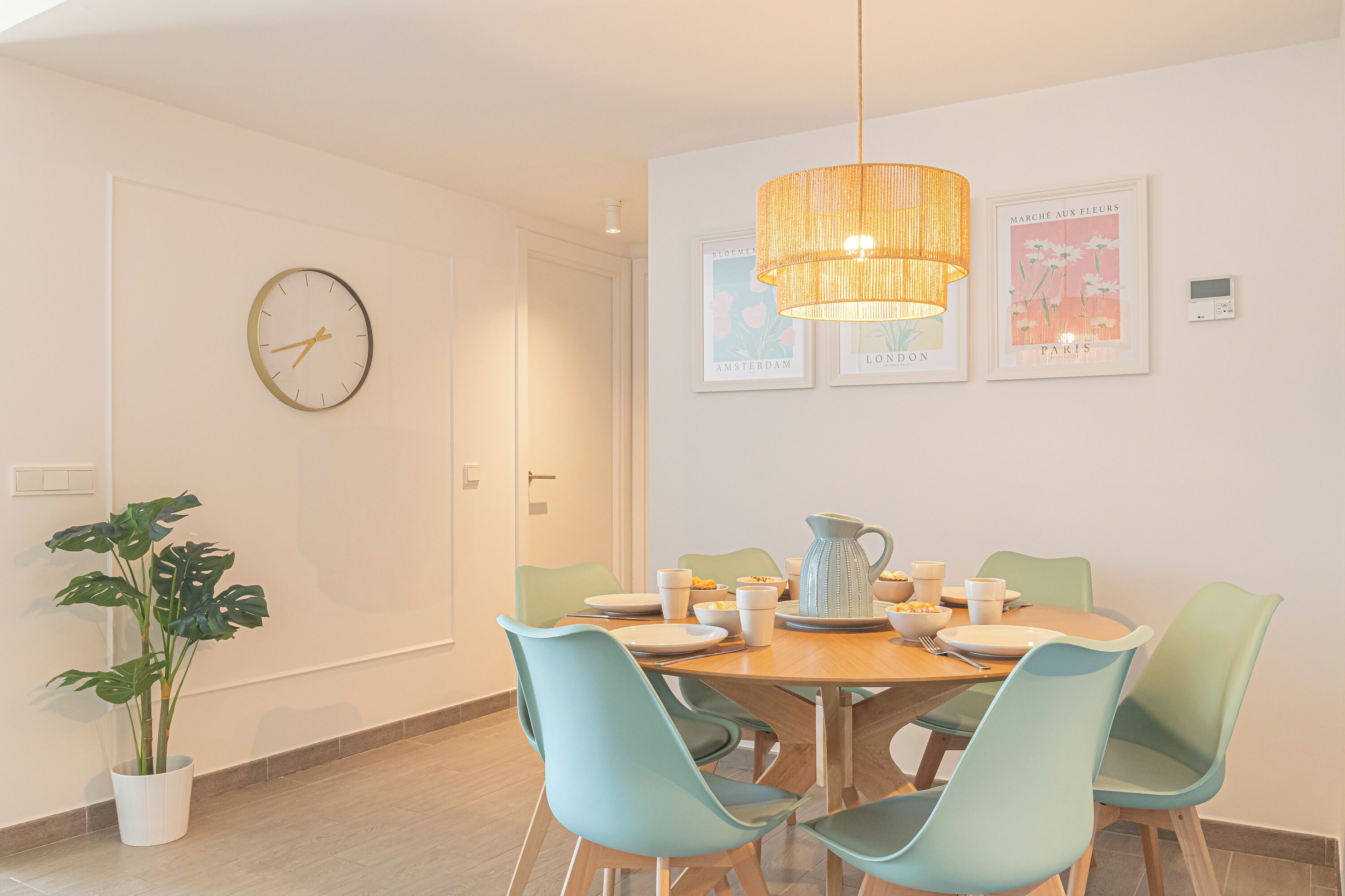 Un comedor con una mesa de madera, cuatro sillas, una planta en maceta, un reloj de pared y cuadros enmarcados.