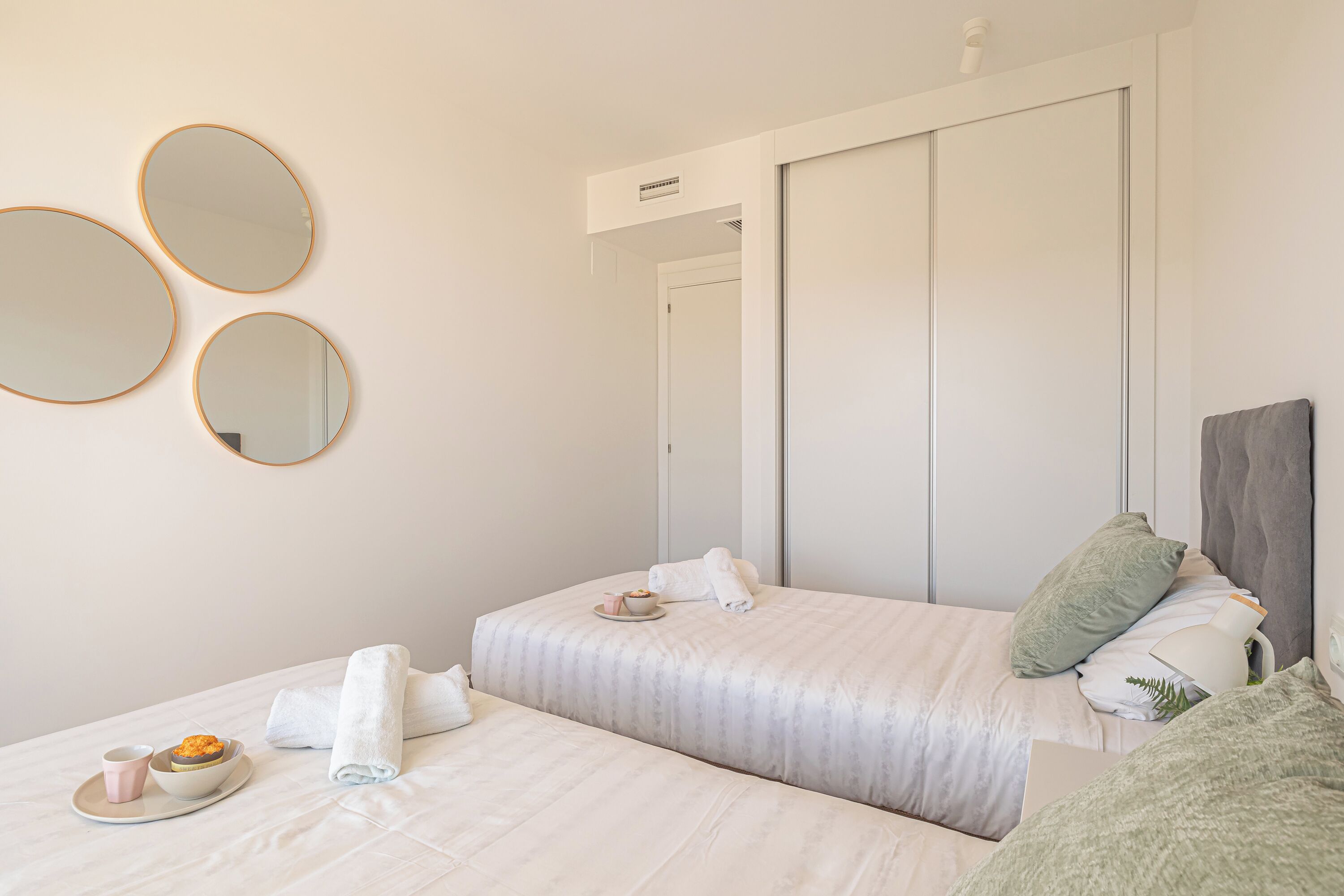 A hotel room with two beds, a large mirror, a bedside table with a lamp, and a small table with a bowl of food.