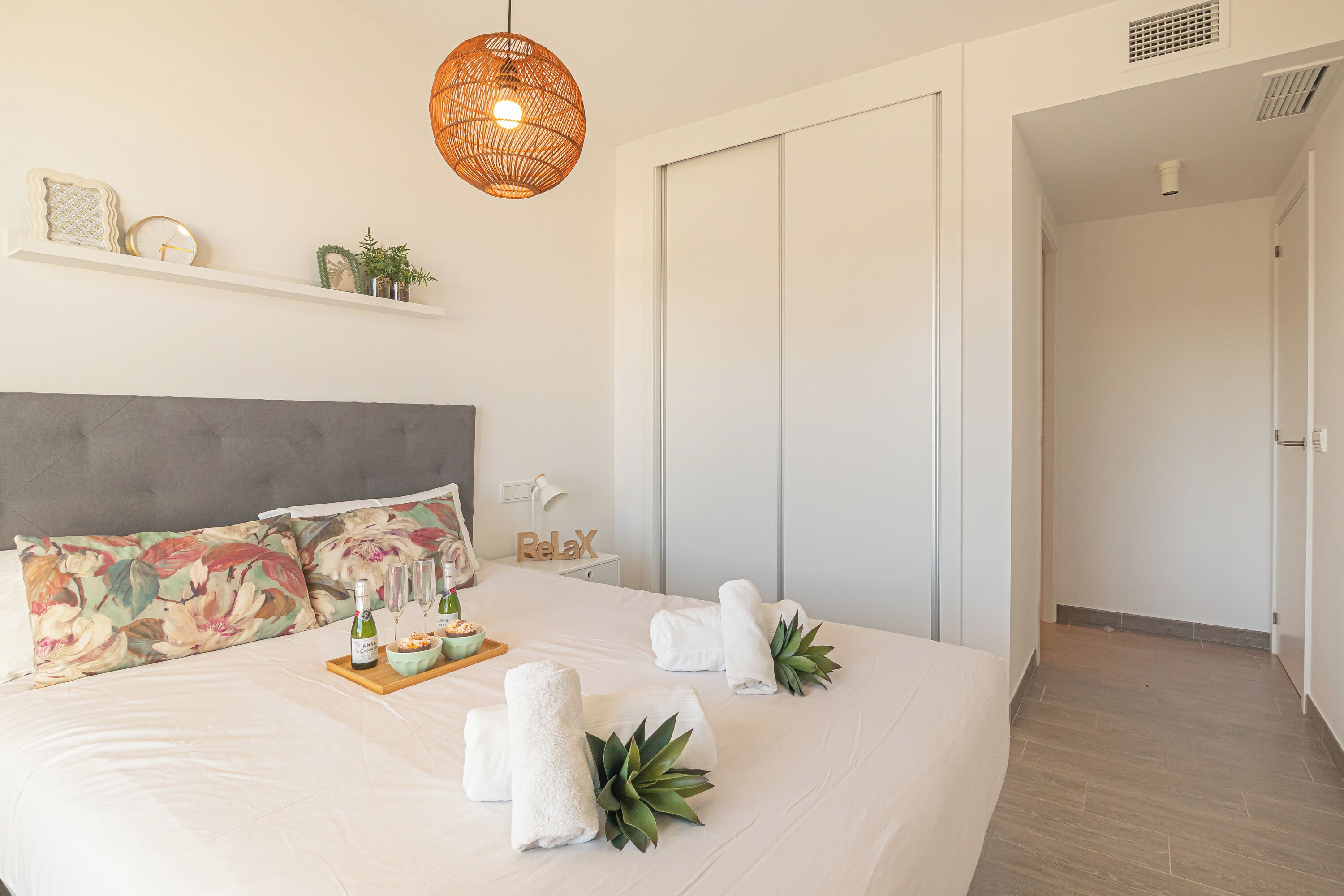 A modern hotel room with a large bed, a headboard, a bedside table with a lamp, a shelf with decorative items, and a door leading to another room.