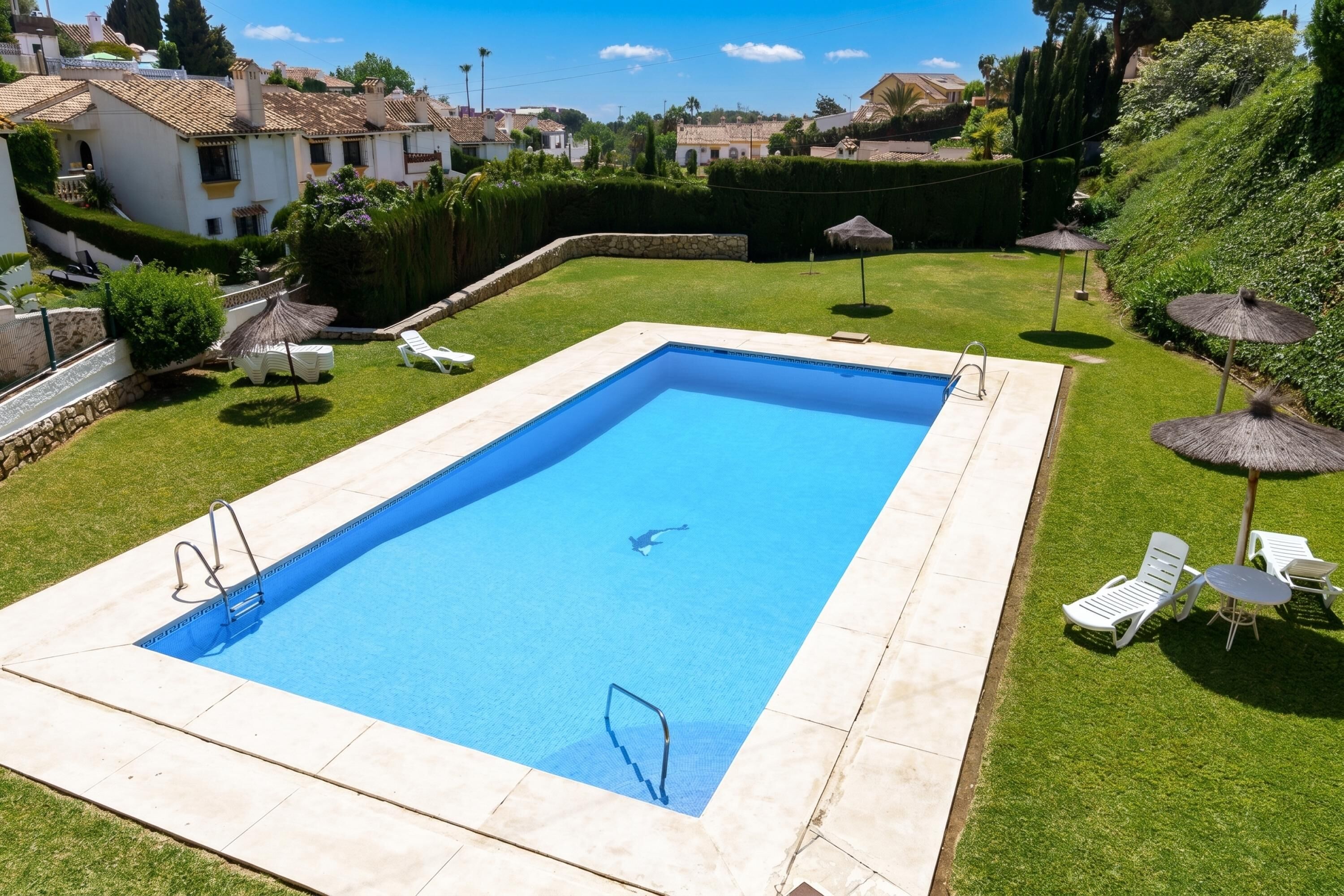 A rectangular swimming pool with a diving board, surrounded by a well-maintained lawn, white lounge chairs, and umbrellas.