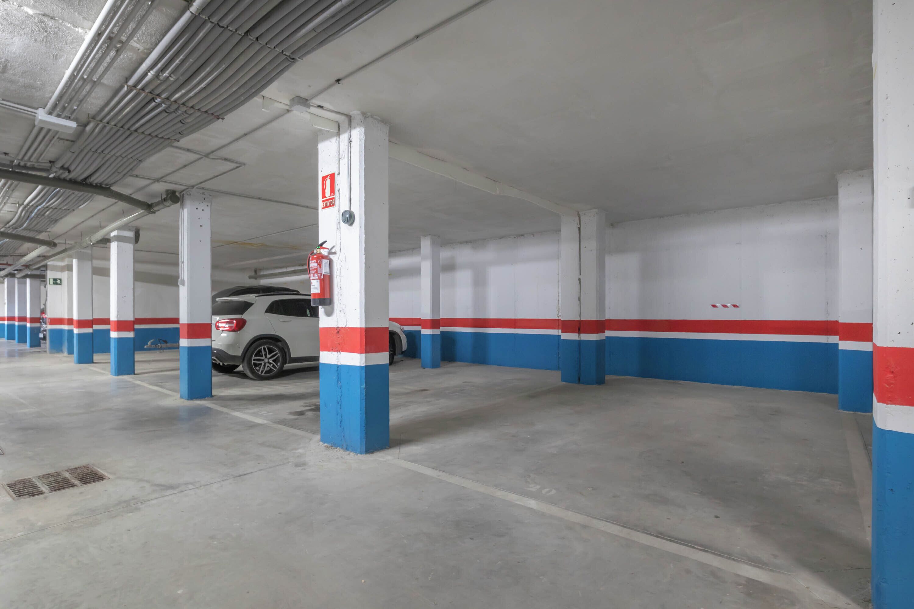 A parking garage with white pillars painted in red and blue stripes, a fire extinguisher mounted on a pillar, and a white car parked in the distance.