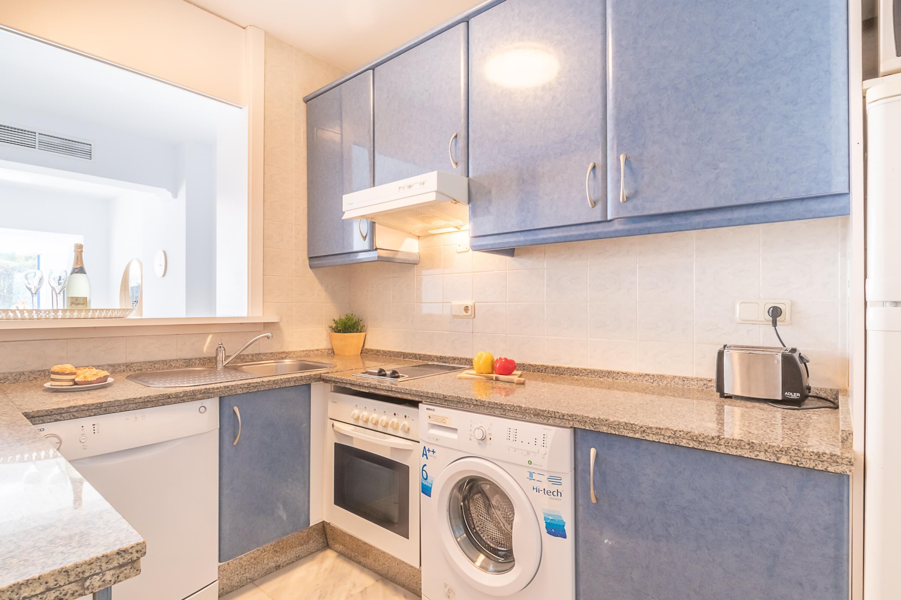 Een compacte keuken met blauwe kastjes, een inbouwoven, een vaatwasser en een wasmachine.