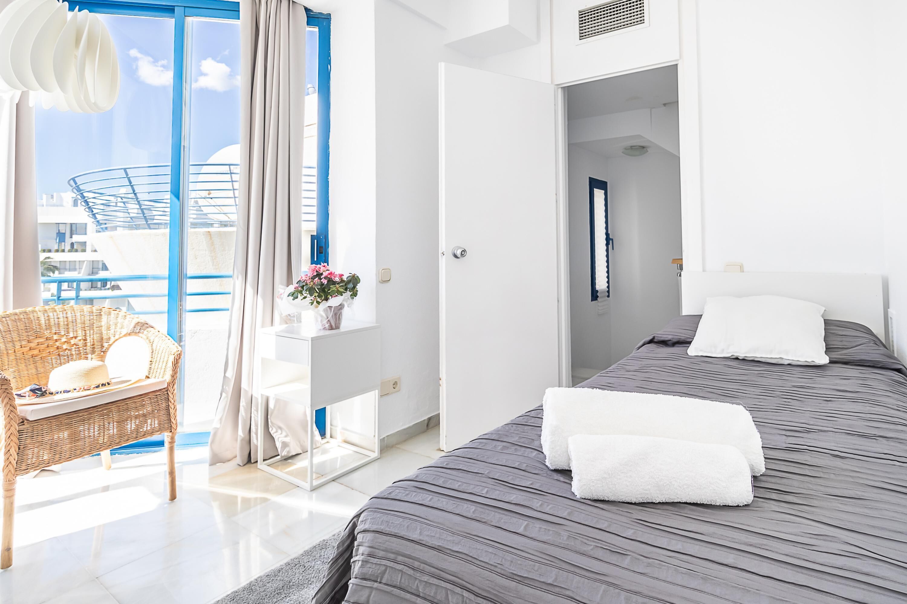Habitación de hotel con una cama grande, una silla de mimbre, una mesita con un florero y vistas al exterior.
