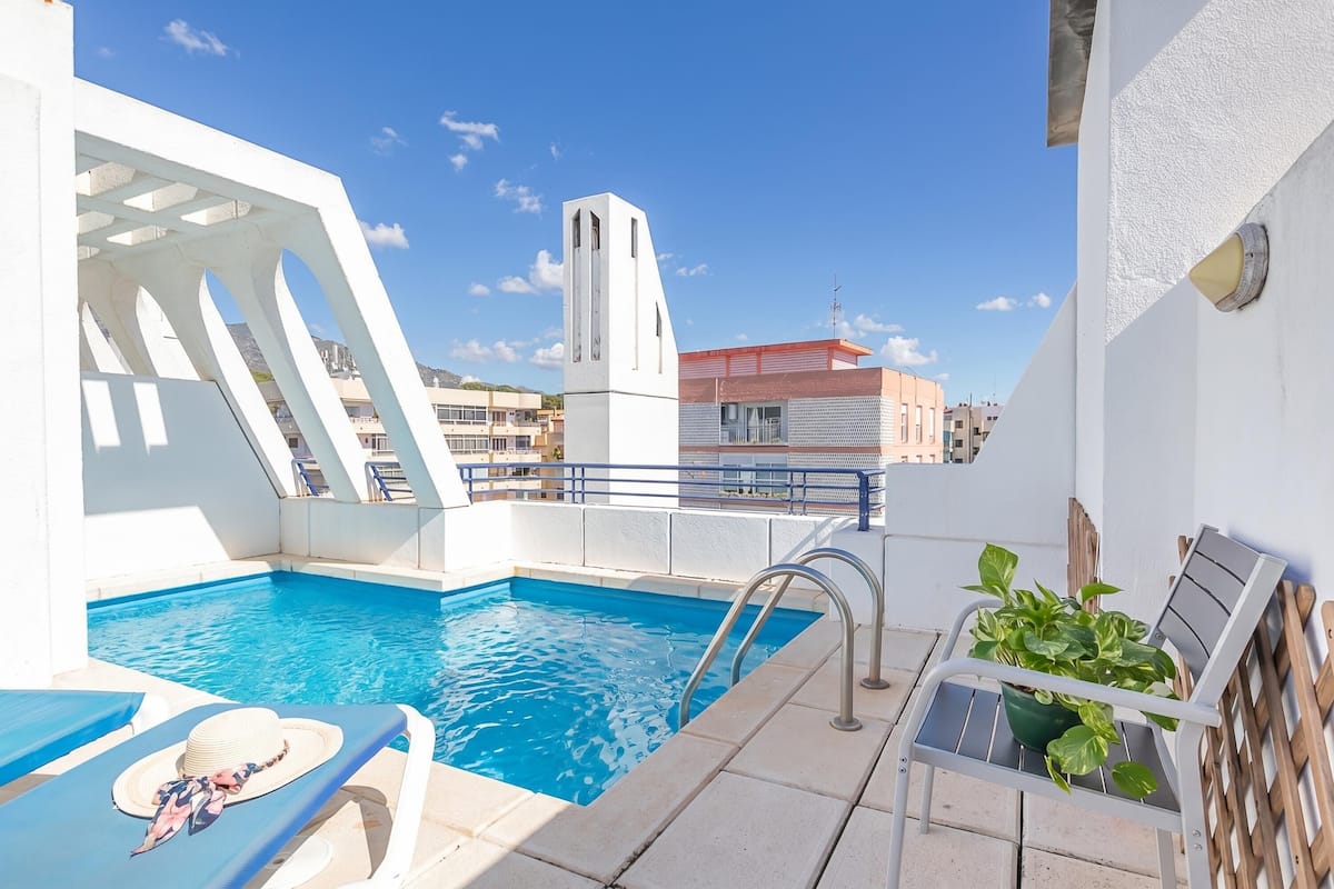 A rooftop pool with a white structure, a chair, and a potted plant.