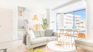 Een lichte woonkamer met een bank, een ronde tafel en uitzicht op een balkon.